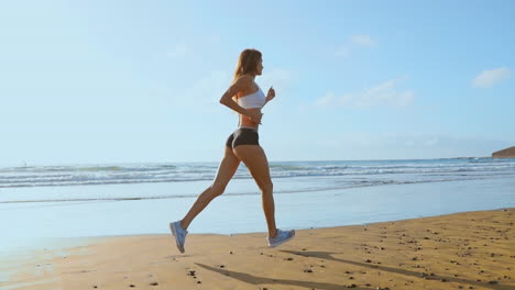 Beautiful-woman-in-sports-shorts-and-t-shirt-running-on-the-beach-with-white-sand-and-blue-ocean-water-on-the-island-in-slow-motion.-Waves-and-sand-hills-on-the-back-won